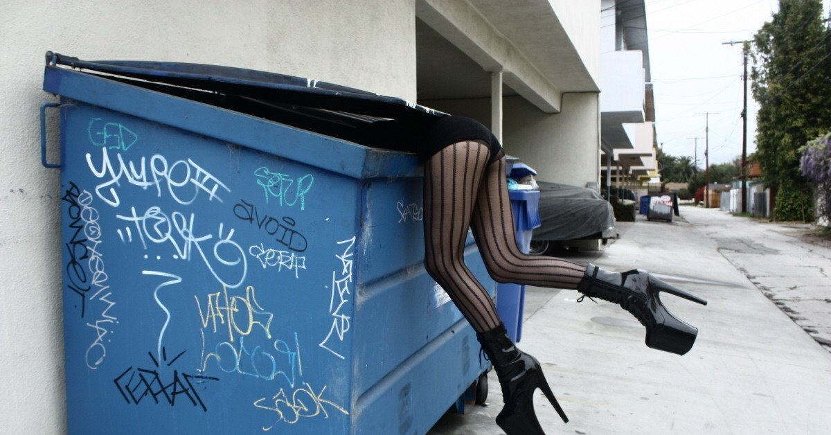 Транс срет. Ноги торчат из мусорного бака. Женщина роется в мусорке.
